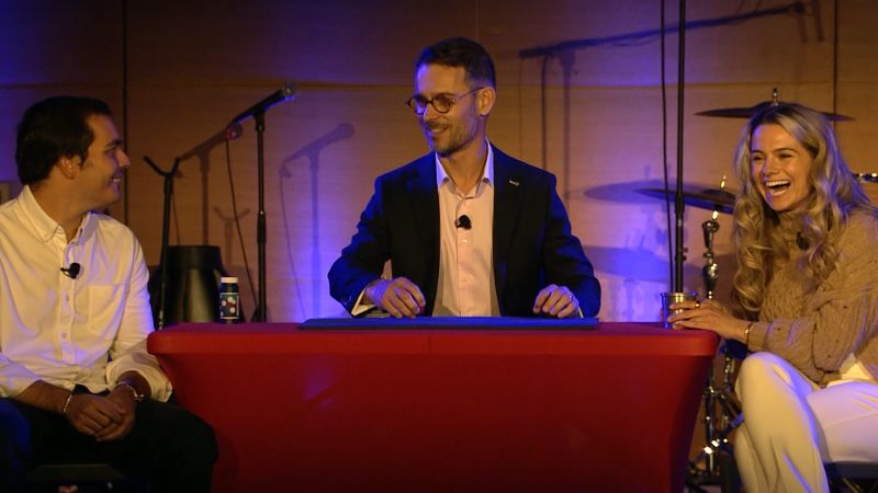 Three characters seated around a small table. FX the magician seats behind the table while both participants on each side are laughing.