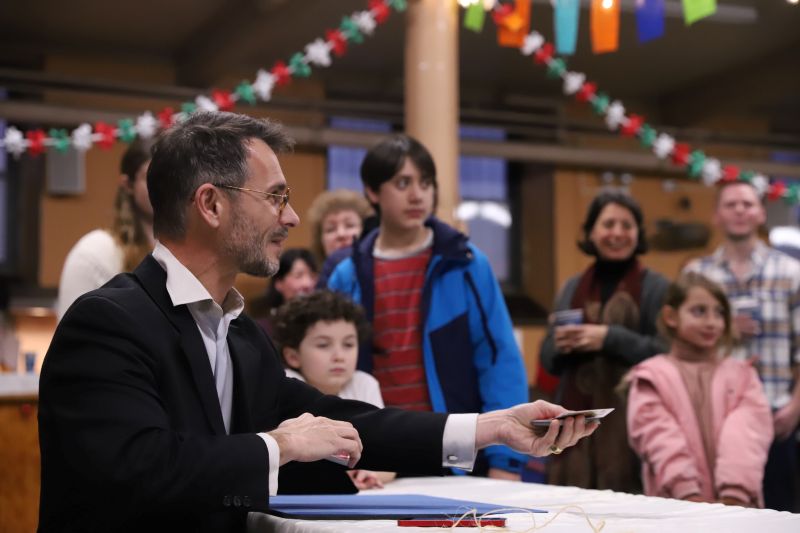 FX the magician seated at a table extends his left hand so a participant can pick a card