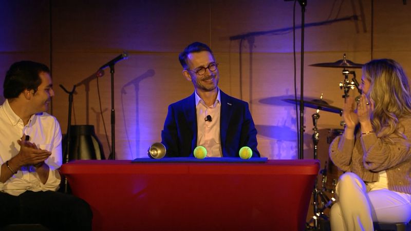 Three characters seated around a small table. FX the magician seats behind the table while both participants on each side are clapping. All have a large smile on their faces. There are 2 tennis balls on the table with an empty cup.