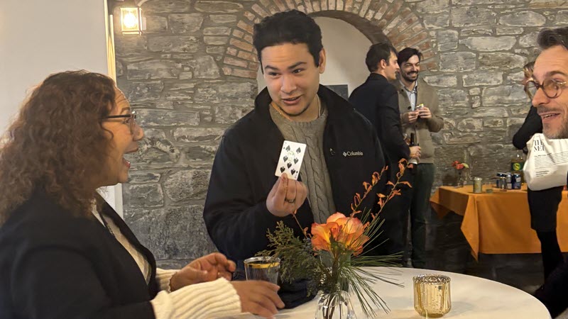 Two participants around a high-top table at a party. Lady on the left is amazed when her son in the center reveals the 7 of spades. FX the magician, is on the right.
